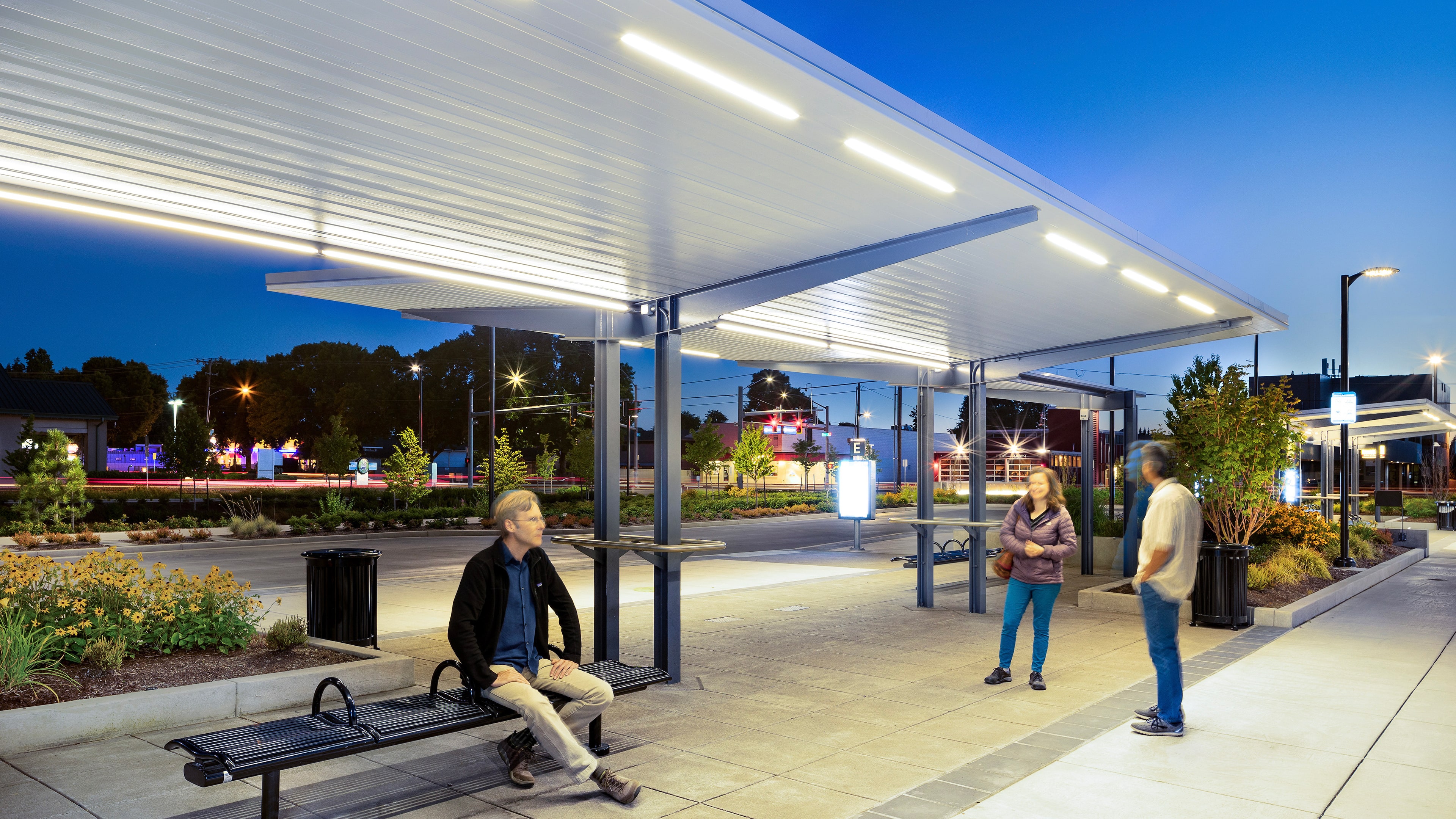 Three people wait at a modern, well-lit outdoor transit station in the evening; one sits on a bench while two stand and talk.