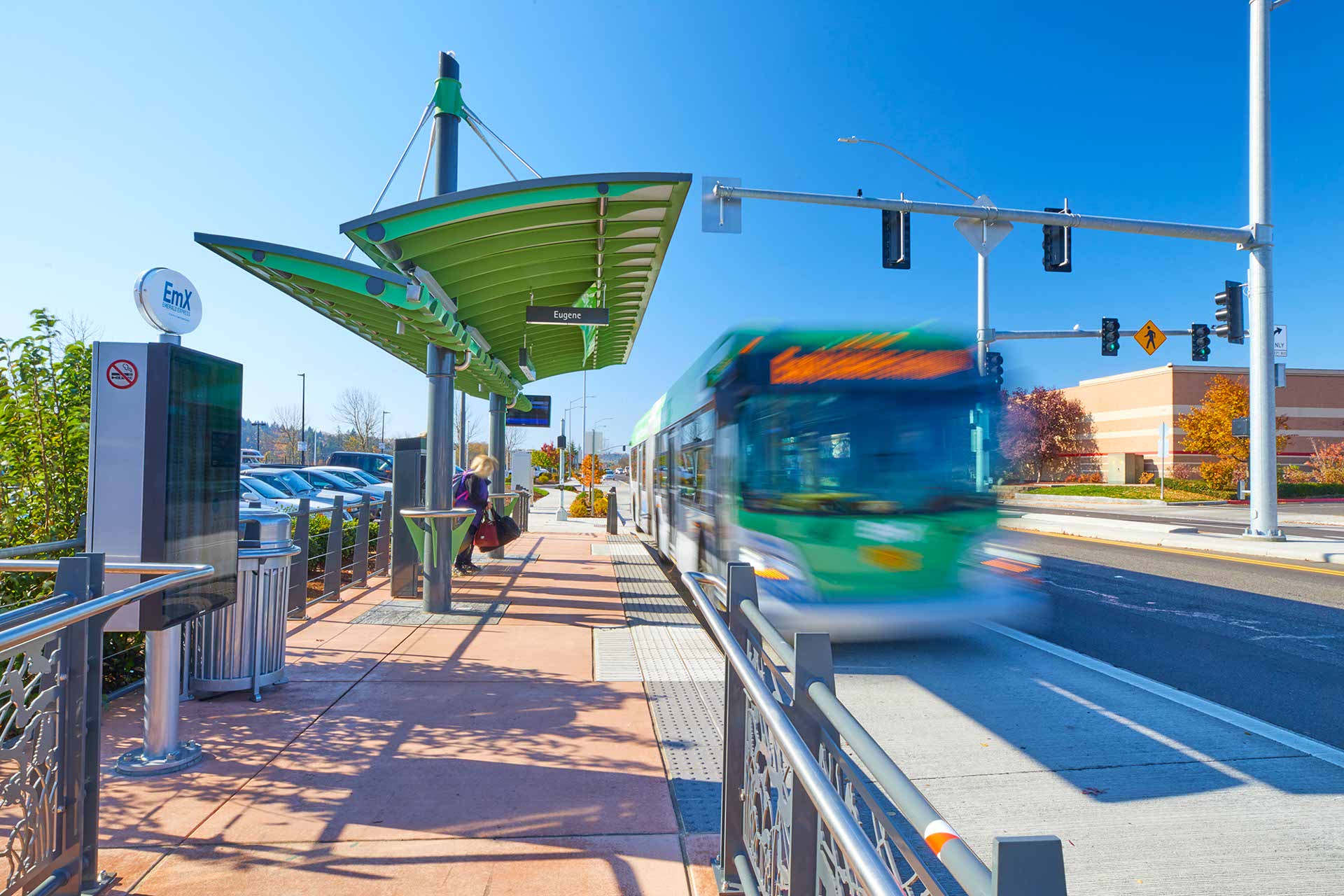 A green and white city bus approaches a modern outdoor bus stop with a green canopy on a sunny day; people wait on the sidewalk.