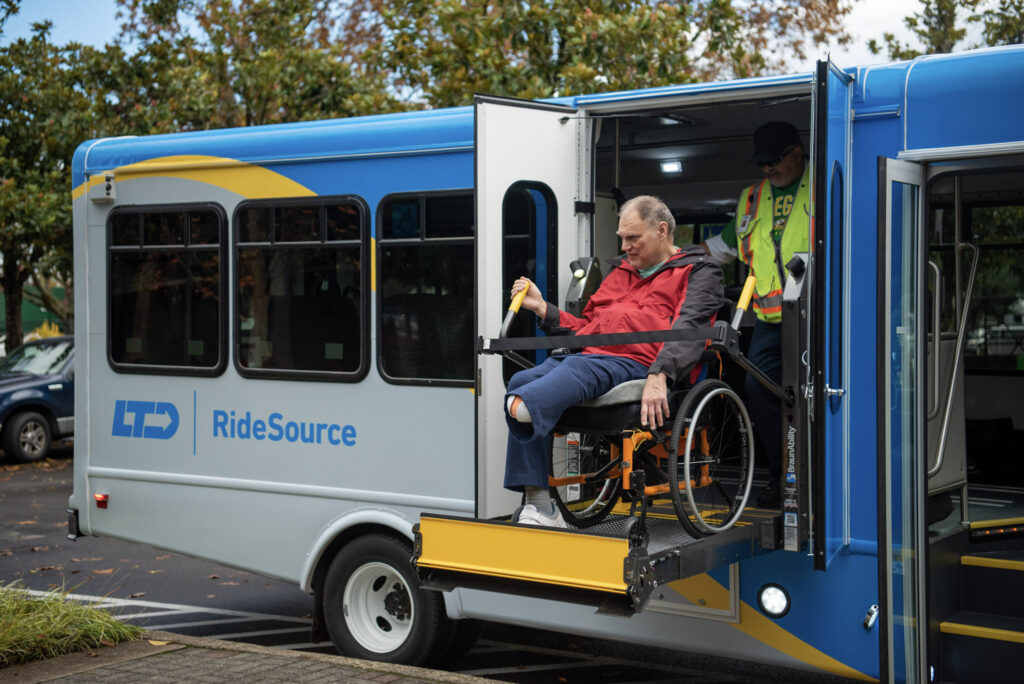 A man in a wheelchair is being lowered from a RideSource accessible van using a lift, assisted by a staff member in a safety vest.