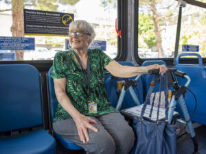 An older woman with glasses sits on a bus, holding a walker and a blue handbag, smiling. Signs for wheelchair seating are visible in the background.