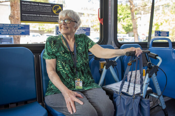 An older woman with glasses sits on a bus, holding a walker and a blue handbag, smiling. Signs for wheelchair seating are visible in the background.