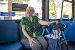 An older woman sits on a bus bench seat, holding a walker and a blue bag, surrounded by signs designating the wheelchair seating area.