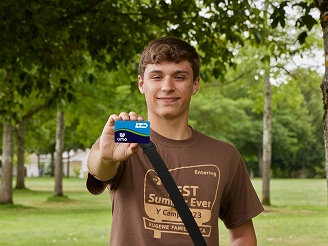product2 A young man stands outdoors holding a blue and white debit card toward the camera, with trees and grass in the background.