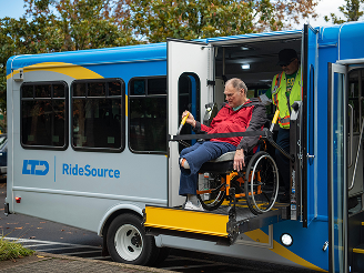 product3 A man in a wheelchair is being assisted by an attendant as he exits a RideSource bus via a wheelchair lift.
