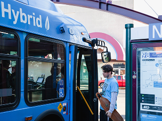 product4 A person holding a longboard waits to board a blue hybrid bus at a city bus stop with a route map visible.