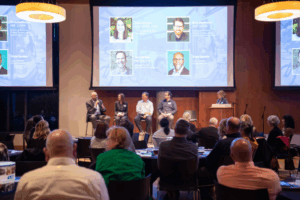 A panel of five people speak on stage in front of an audience, with their names and photos displayed on large screens behind them.