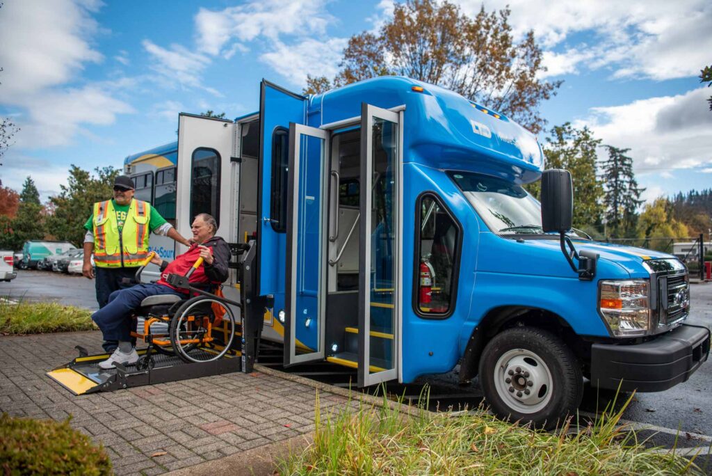 A person in a wheelchair is assisted by a worker as they board a blue accessible shuttle bus using a wheelchair lift on a cloudy day.