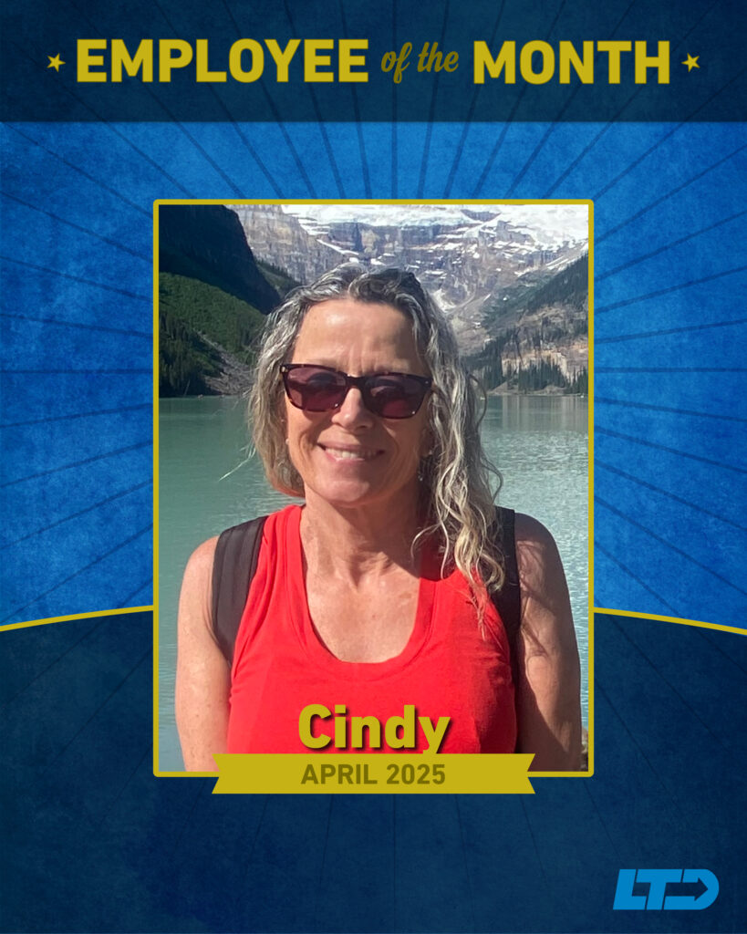 A woman with wavy hair and sunglasses smiles outdoors by a lake and mountains, featured as "Employee of the Month" for April 2025, named Cindy.