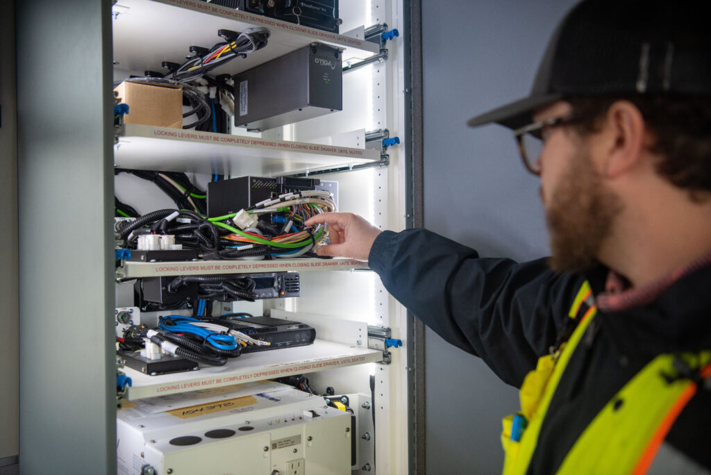 A person wearing a safety vest inspects and arranges cables inside an open electrical equipment cabinet.