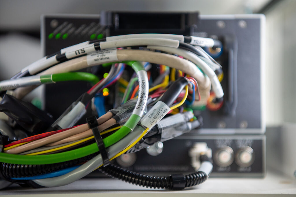Close-up of a bundle of labeled cables connected to an electronic device with indicator lights and two circular ports visible in the background.