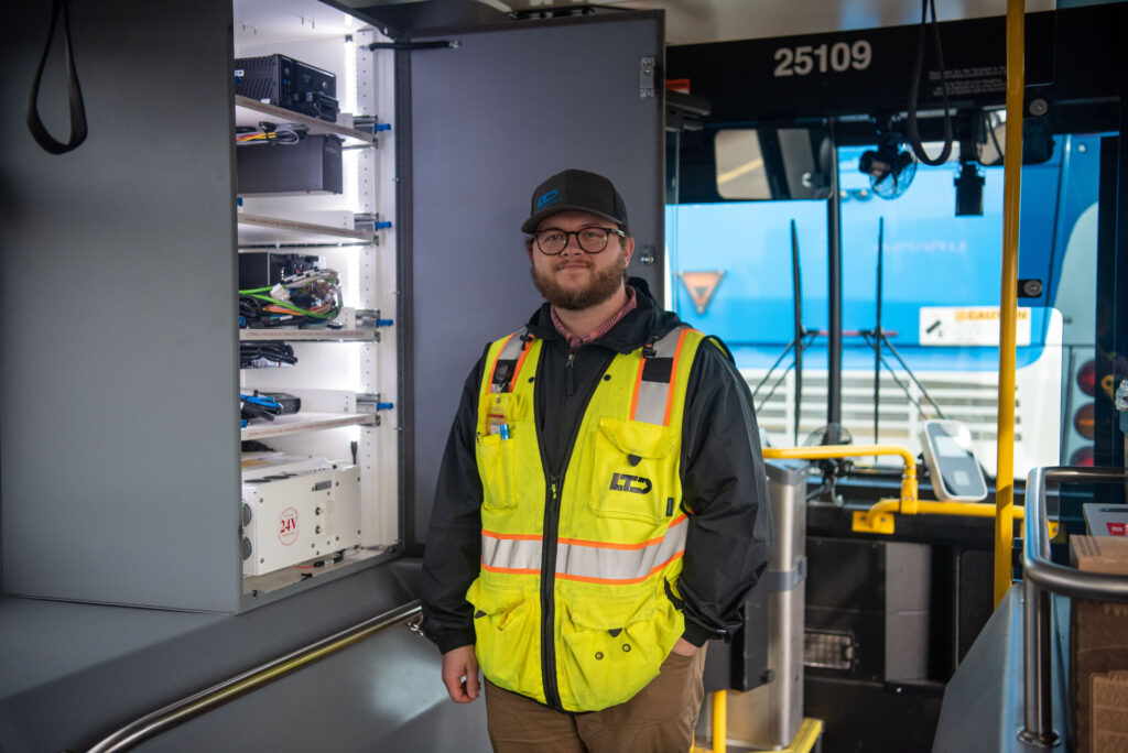 A person in a safety vest and hat stands inside a bus next to an open electrical cabinet with visible wires and equipment.
