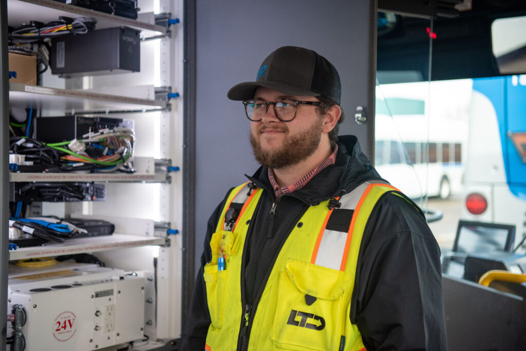 A man wearing glasses, a hat, and a high-visibility jacket stands next to electronic equipment and wiring inside a vehicle.