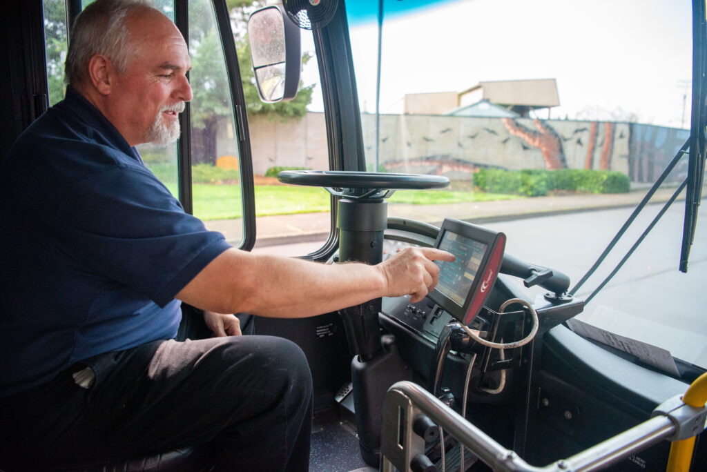 A man in a blue shirt sits in a bus driver's seat and interacts with a touchscreen device mounted next to the steering wheel.