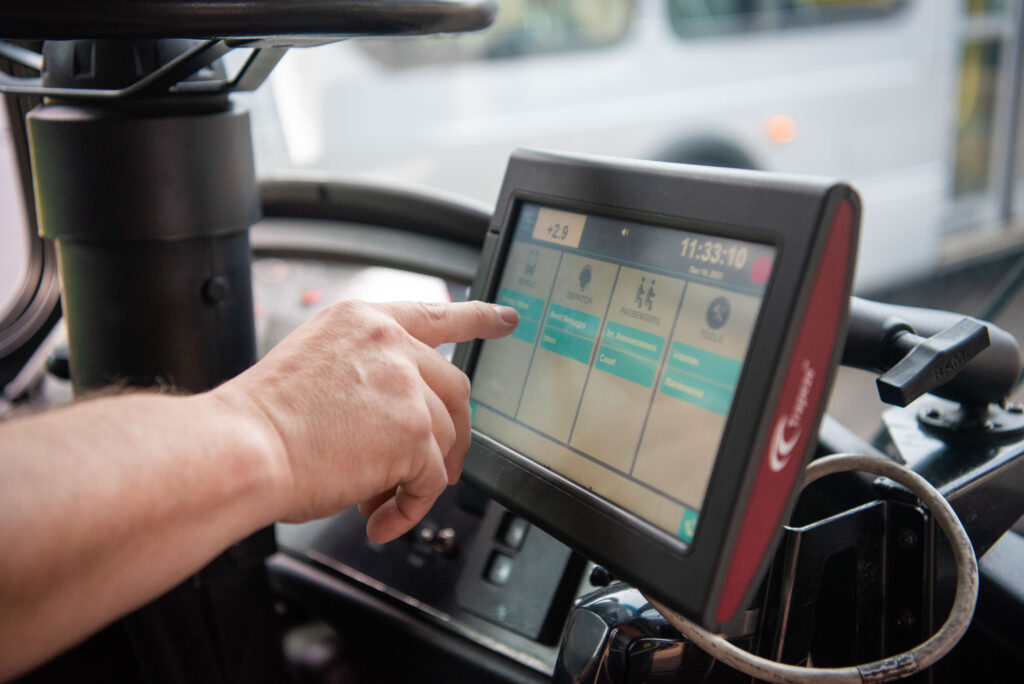 A person operates a touchscreen control panel inside a vehicle, with a bus visible through the window in the background.