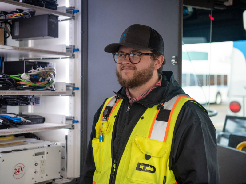 A man wearing a yellow safety vest and black cap stands beside electrical equipment racks inside a vehicle.