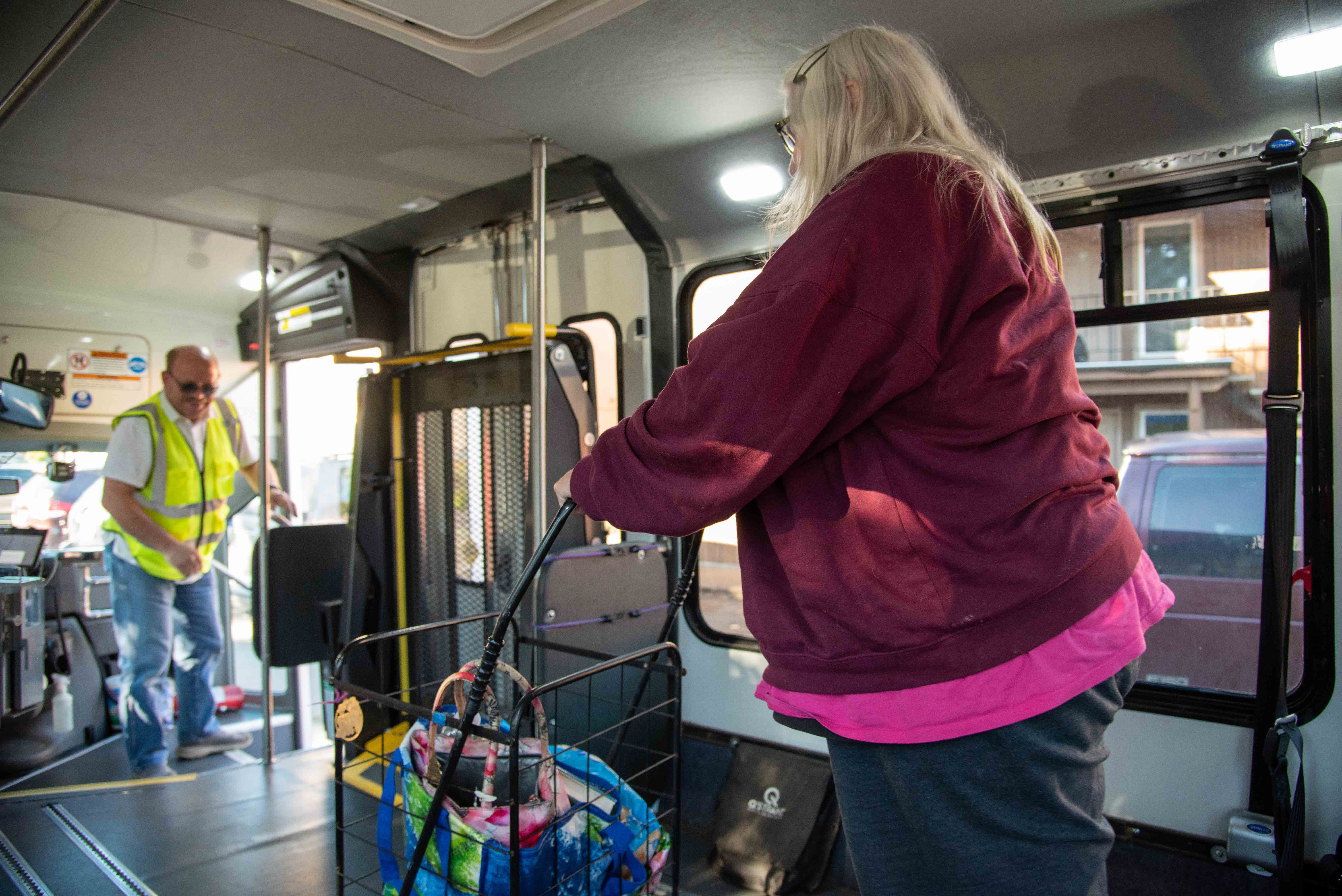 A woman with a shopping cart boards a bus while a man in a yellow safety vest stands near the door inside the vehicle.