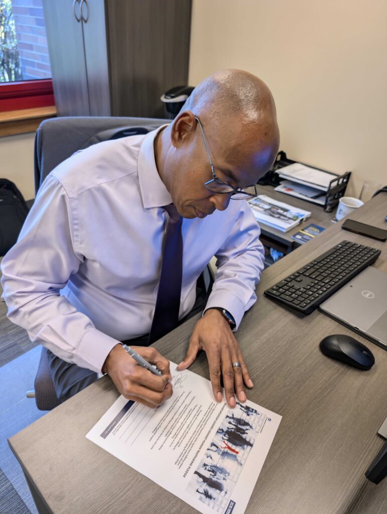 A man in a dress shirt and tie sits at an office desk, signing a printed document with a pen. A computer keyboard, monitor, and office supplies are visible on the desk.