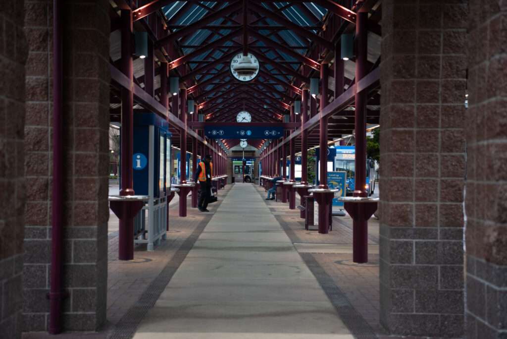 Covered walkway at a transportation terminal with a clock overhead, information signs, and people standing or walking along the path.