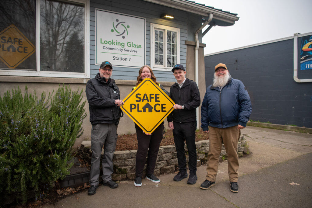 Four adults stand outside Looking Glass Community Services, holding a yellow "Safe Place" sign and posing for a group photo.
