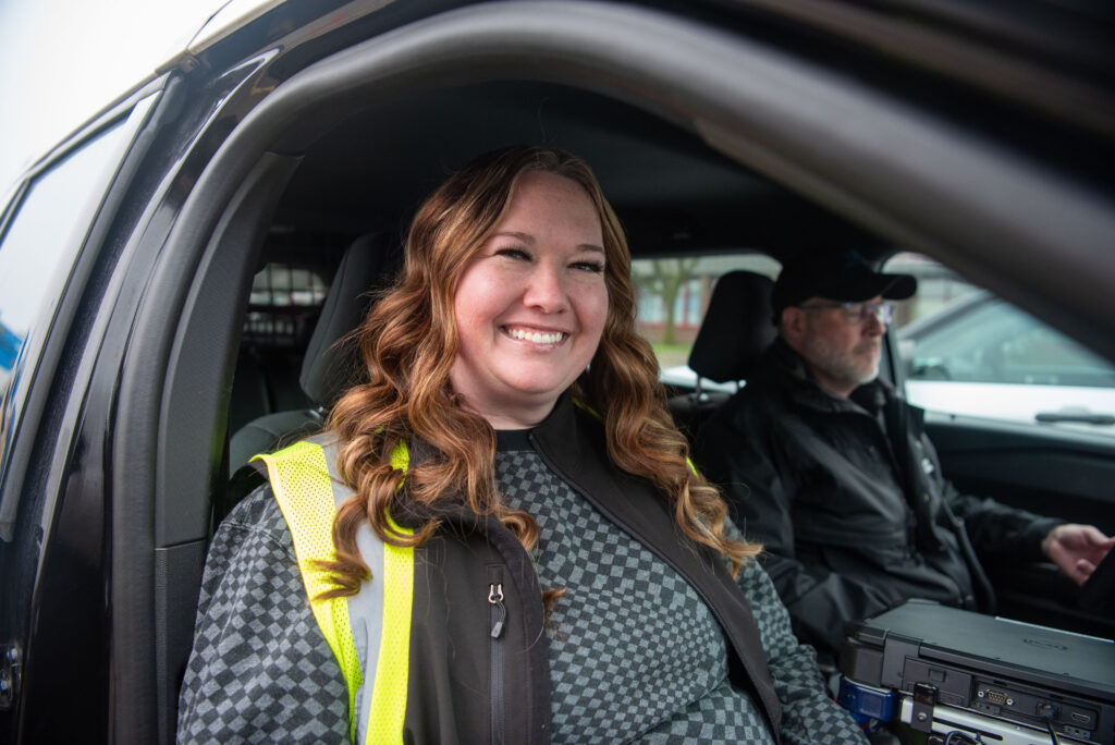 A woman wearing a safety vest smiles while sitting in the front seat of a car, with a man in the driver's seat next to her.