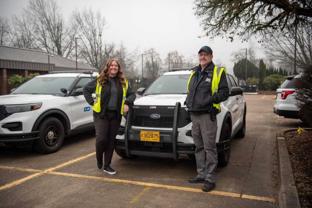 Two people wearing reflective vests stand in front of a white SUV with an Oregon license plate in a parking lot on a cloudy day.