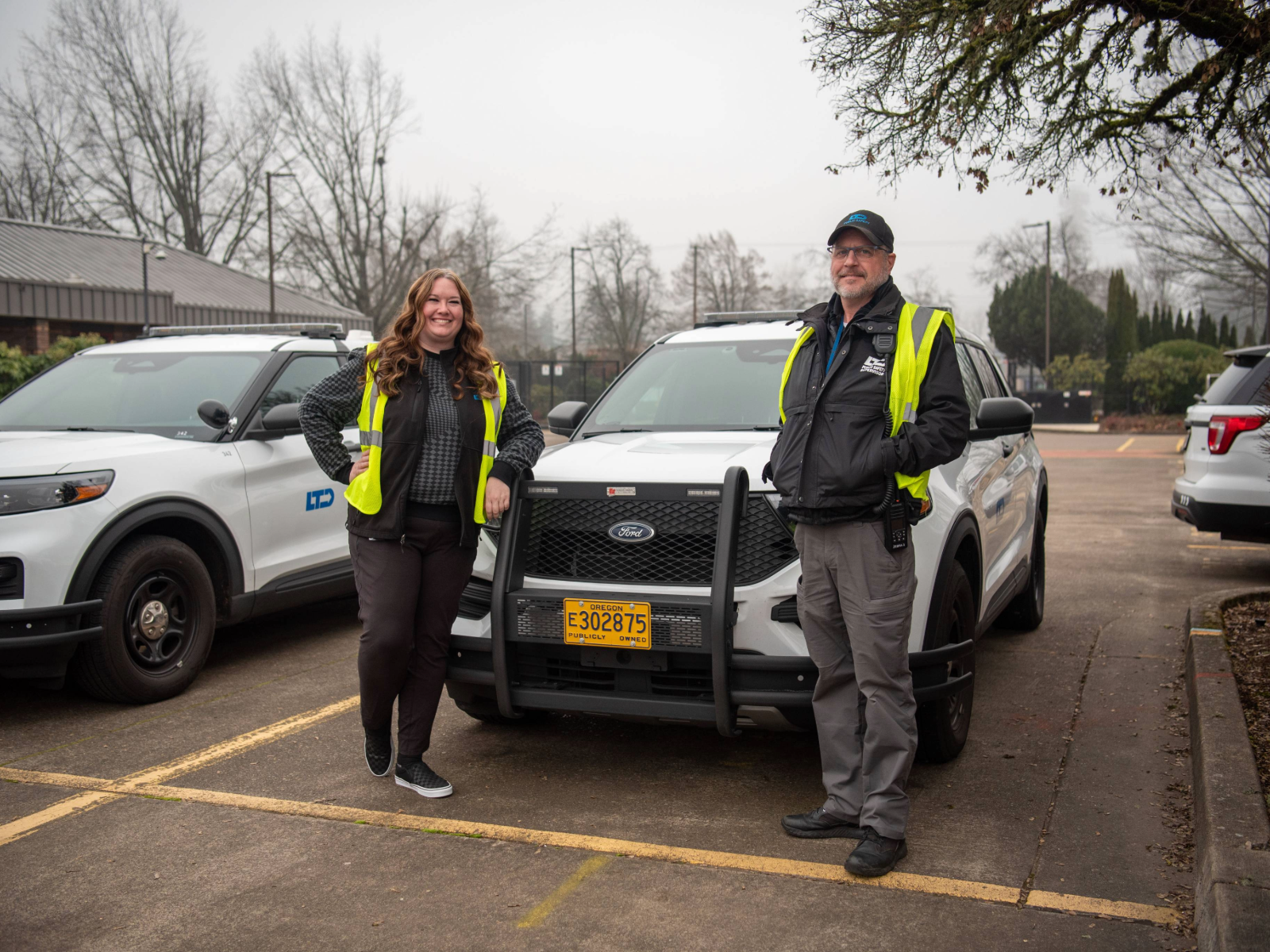 Two people wearing yellow safety vests stand in front of a white SUV with Oregon license plates in a parking lot, with two other similar vehicles parked nearby.