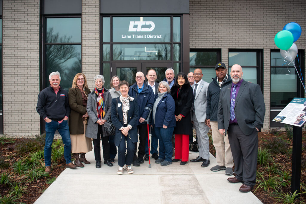 A group of people stand smiling in front of the Lane Transit District building entrance, with balloons and an informational sign visible on the right.
