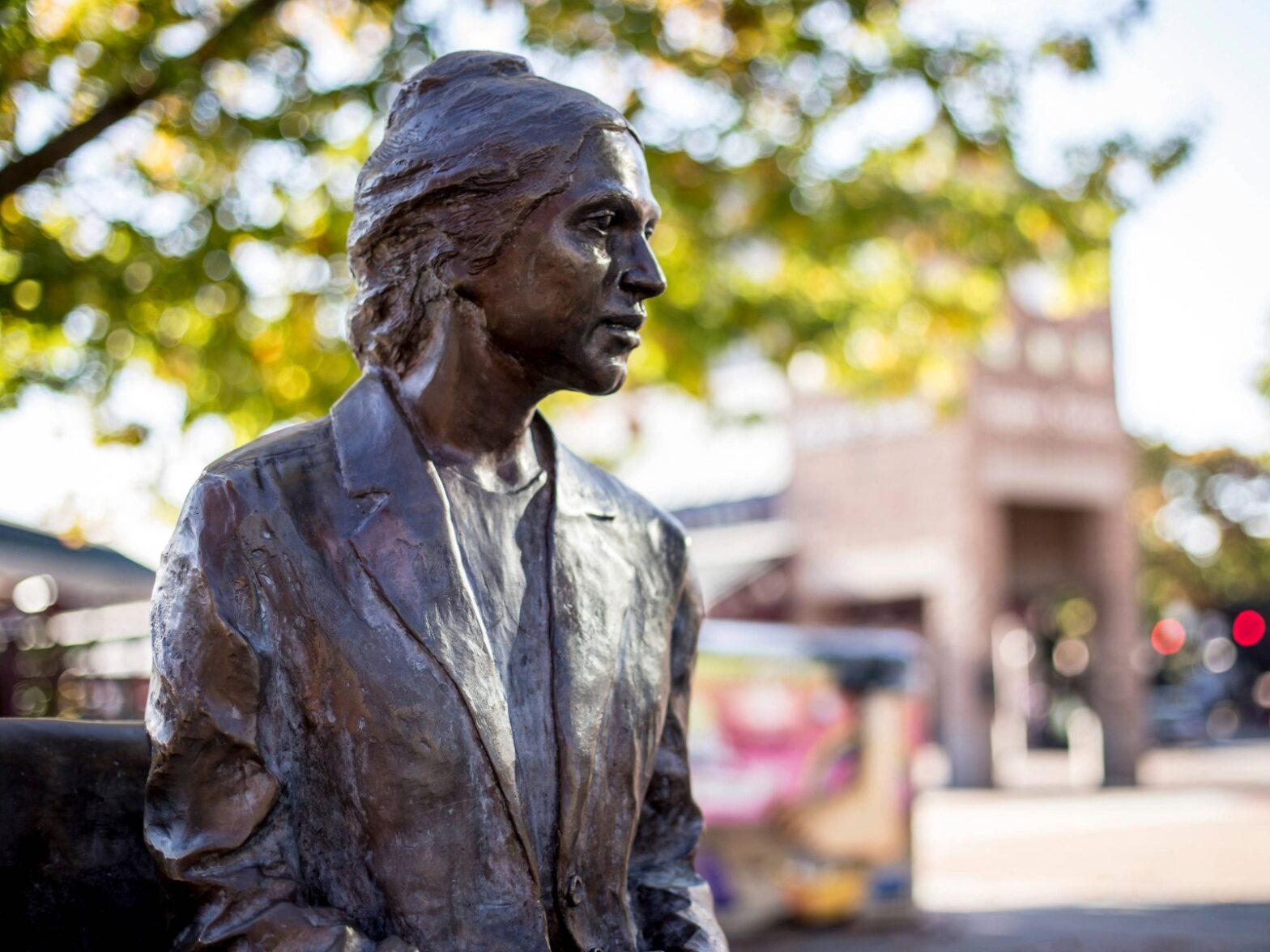 A bronze statue of a seated woman in a blazer, with trees and a building in the blurred background.