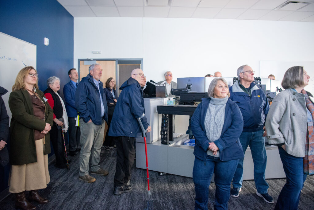 A group of adults stand in a modern office, facing a presentation or screen that is out of view. Some appear attentive, and a few are wearing jackets.