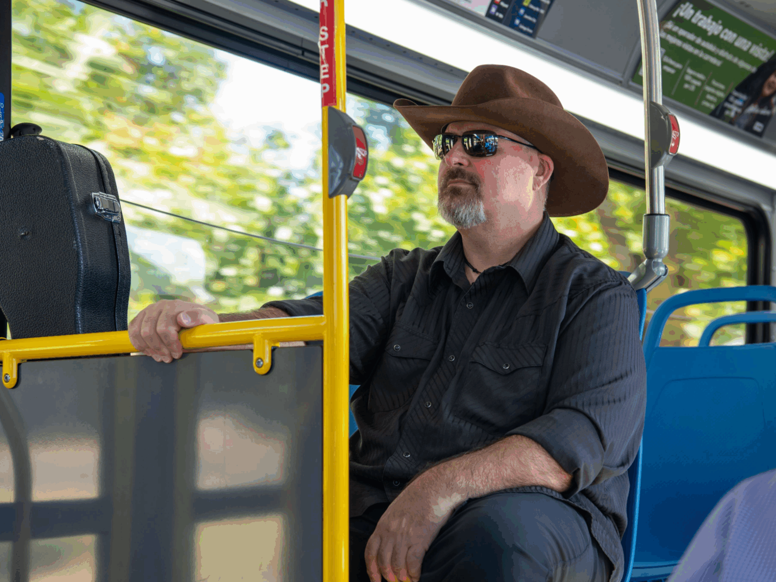 A man wearing a wide-brimmed hat and sunglasses sits on a city bus next to a black guitar case.