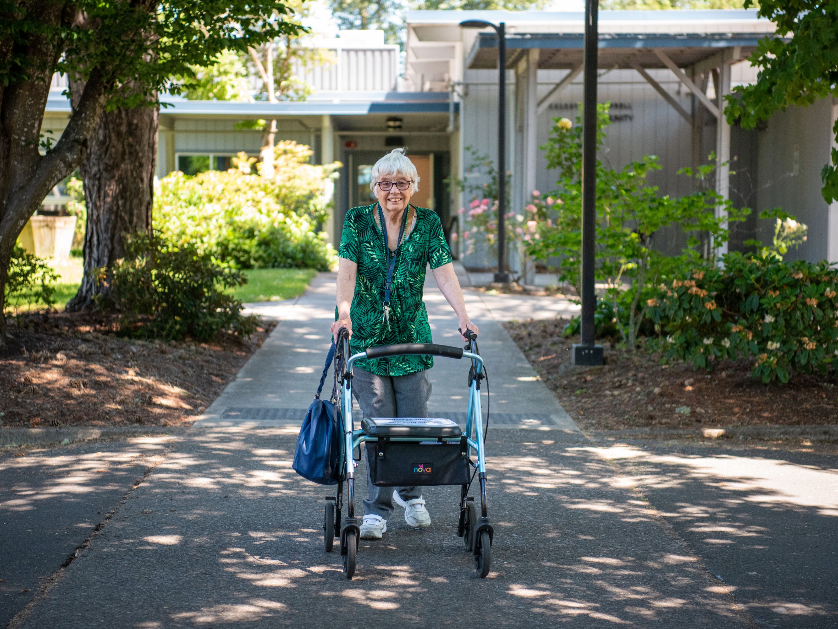 An older woman with gray hair and glasses uses a walker along a paved path outside a building, surrounded by greenery on a sunny day.