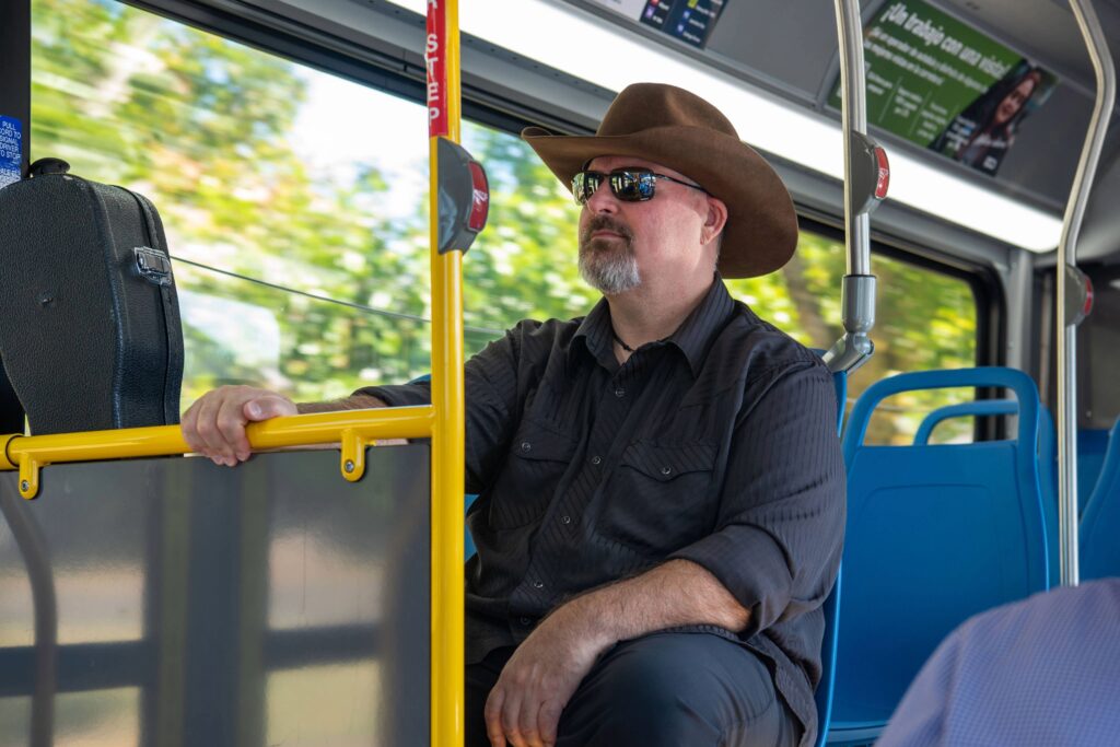 A man wearing a wide-brimmed hat and sunglasses sits on a blue seat inside a city bus, holding a yellow railing.