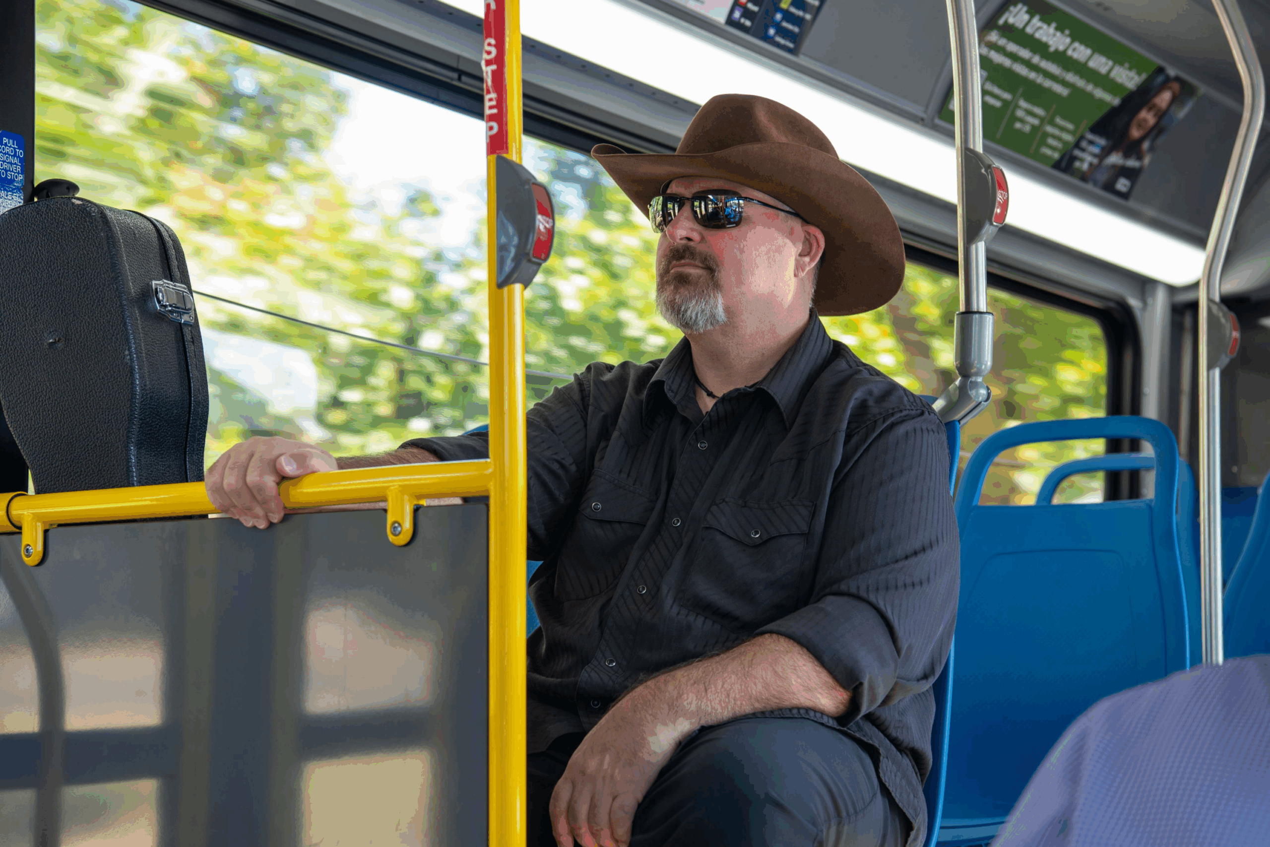A man wearing a wide-brimmed brown hat and sunglasses sits on a bus, holding a yellow rail. A black case rests nearby, and empty blue seats are in the background.