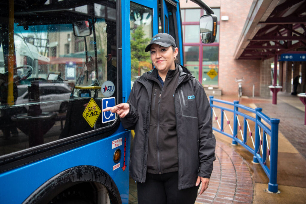 A bus driver stands next to a blue bus, wearing a black jacket and hat, smiling and pointing at the bus door.
