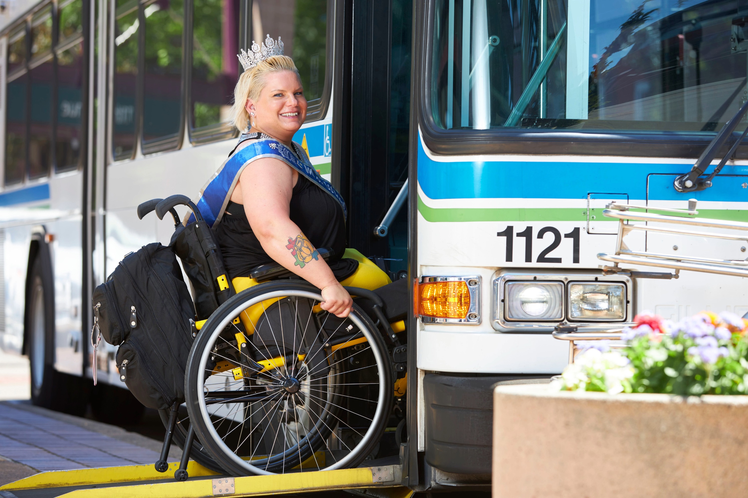 A woman in a wheelchair wearing a sash and crown boards a city bus using a wheelchair ramp on a sunny day.
