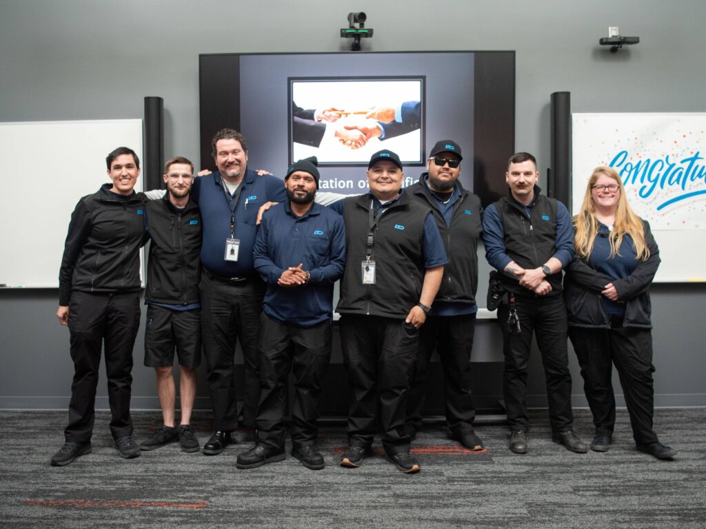 A group of nine people in matching uniforms stand together and smile in front of a presentation screen and congratulatory sign.