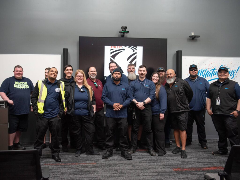 A group of fifteen people pose and smile for a photo in a modern office or training room, standing in front of a screen and wall art.