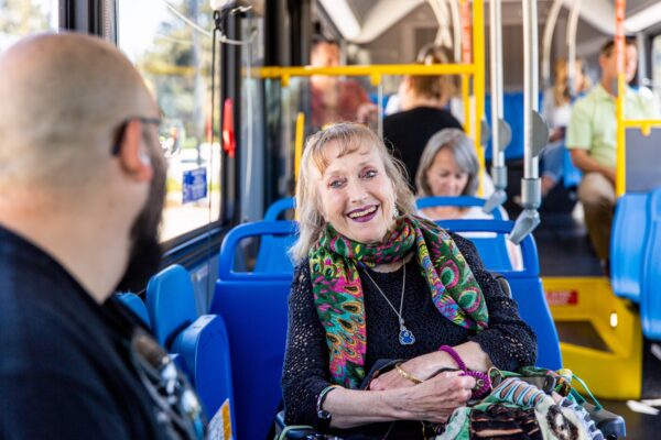 An older woman smiles and talks with a man while sitting on a city bus. Other passengers are seated in the background.
