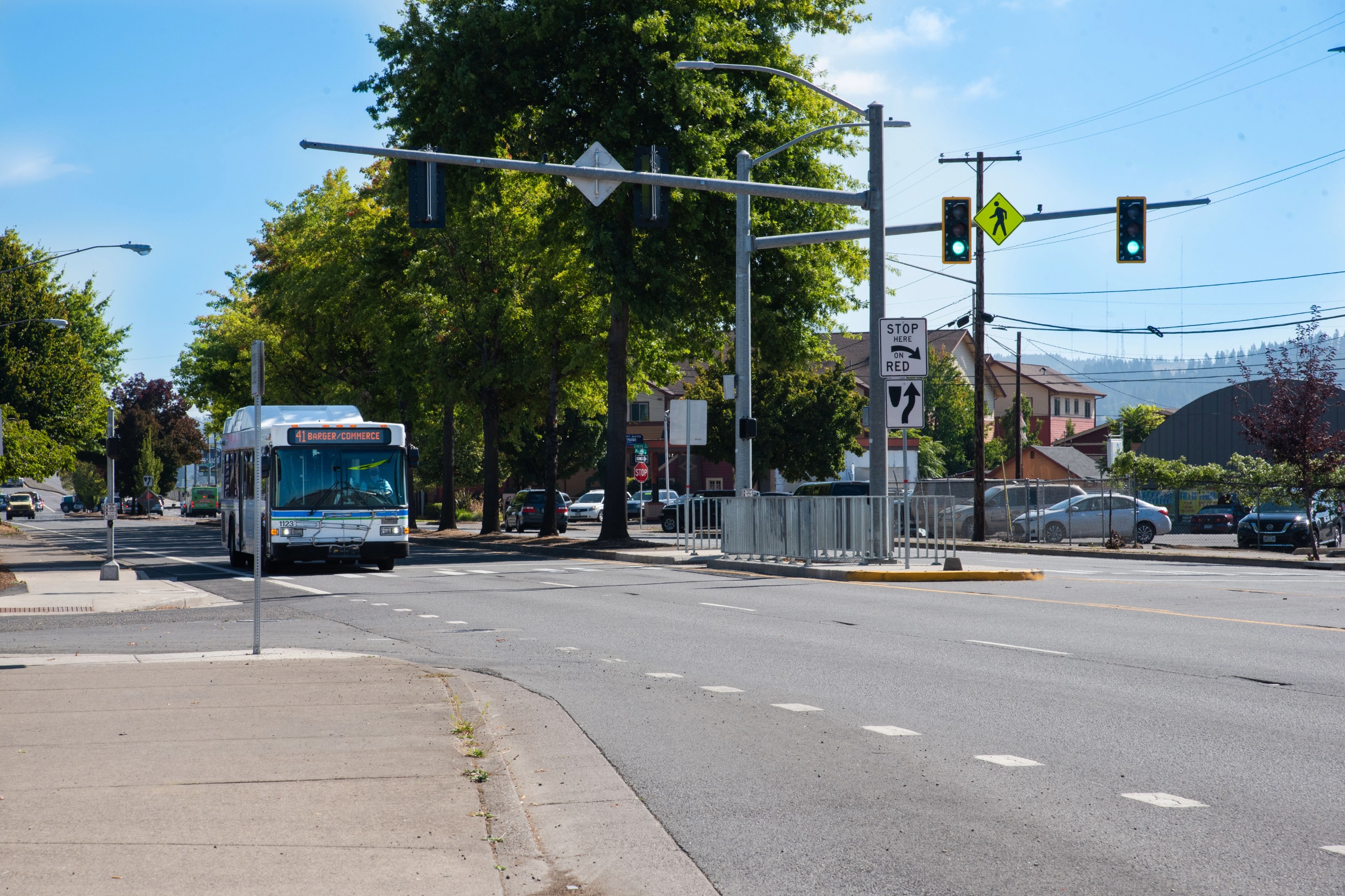 A city bus approaches an intersection with a green traffic light; street signs, crosswalk, and trees line the road on a clear day.