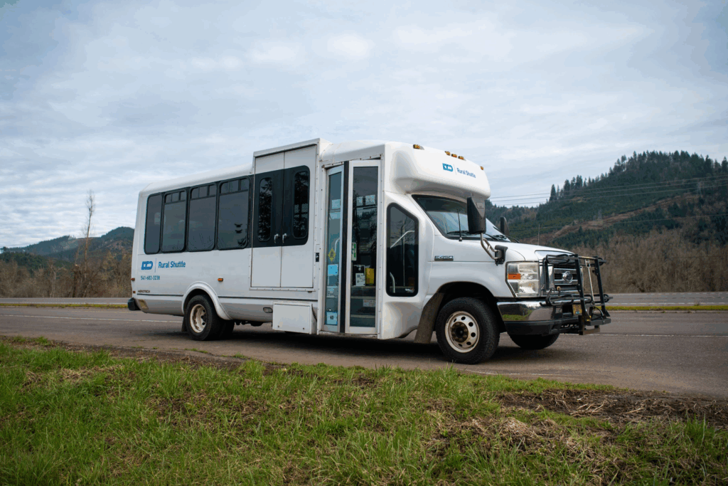 A small white shuttle bus is parked on the side of a rural road with grassy fields and forested hills in the background under a cloudy sky.