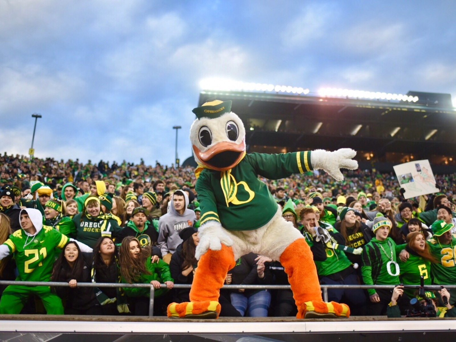 A duck mascot poses in front of a large crowd at a sports stadium, with many fans dressed in green and yellow clothing.