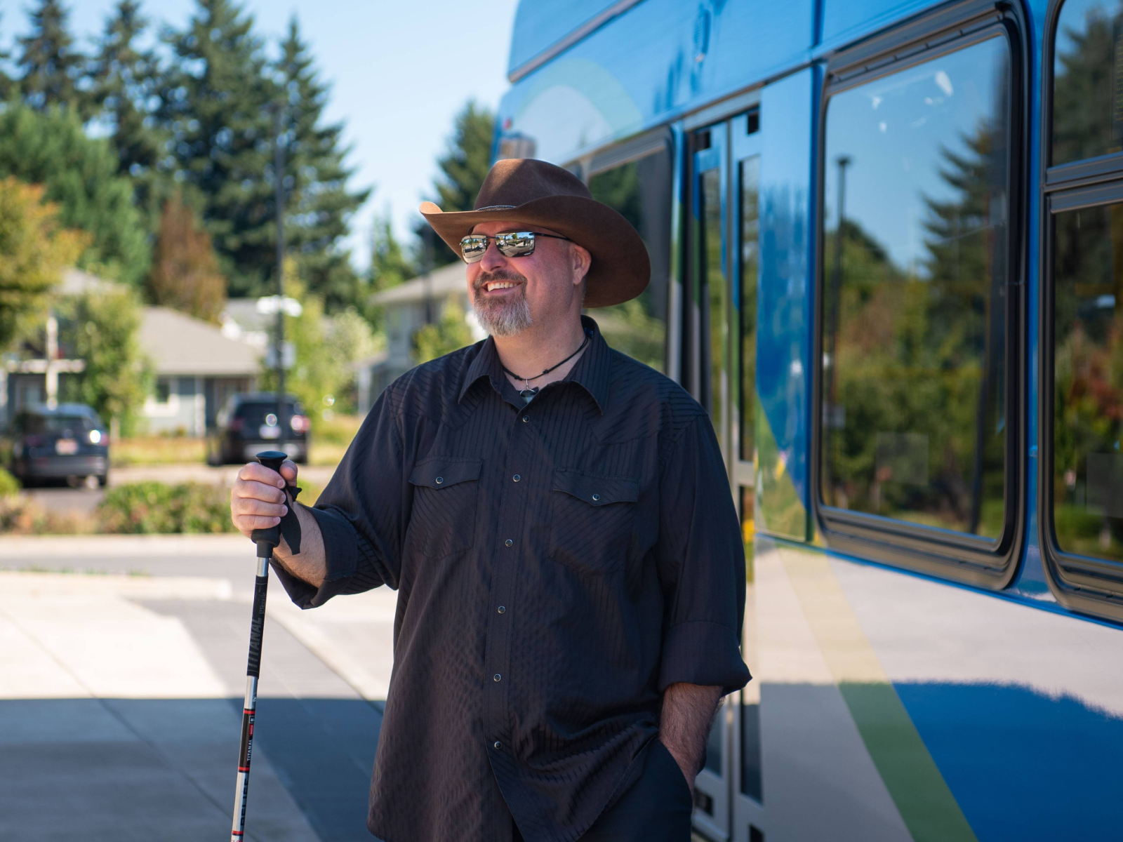 A man in a cowboy hat and sunglasses stands beside a blue bus, holding a mobility cane, with trees and houses in the background.