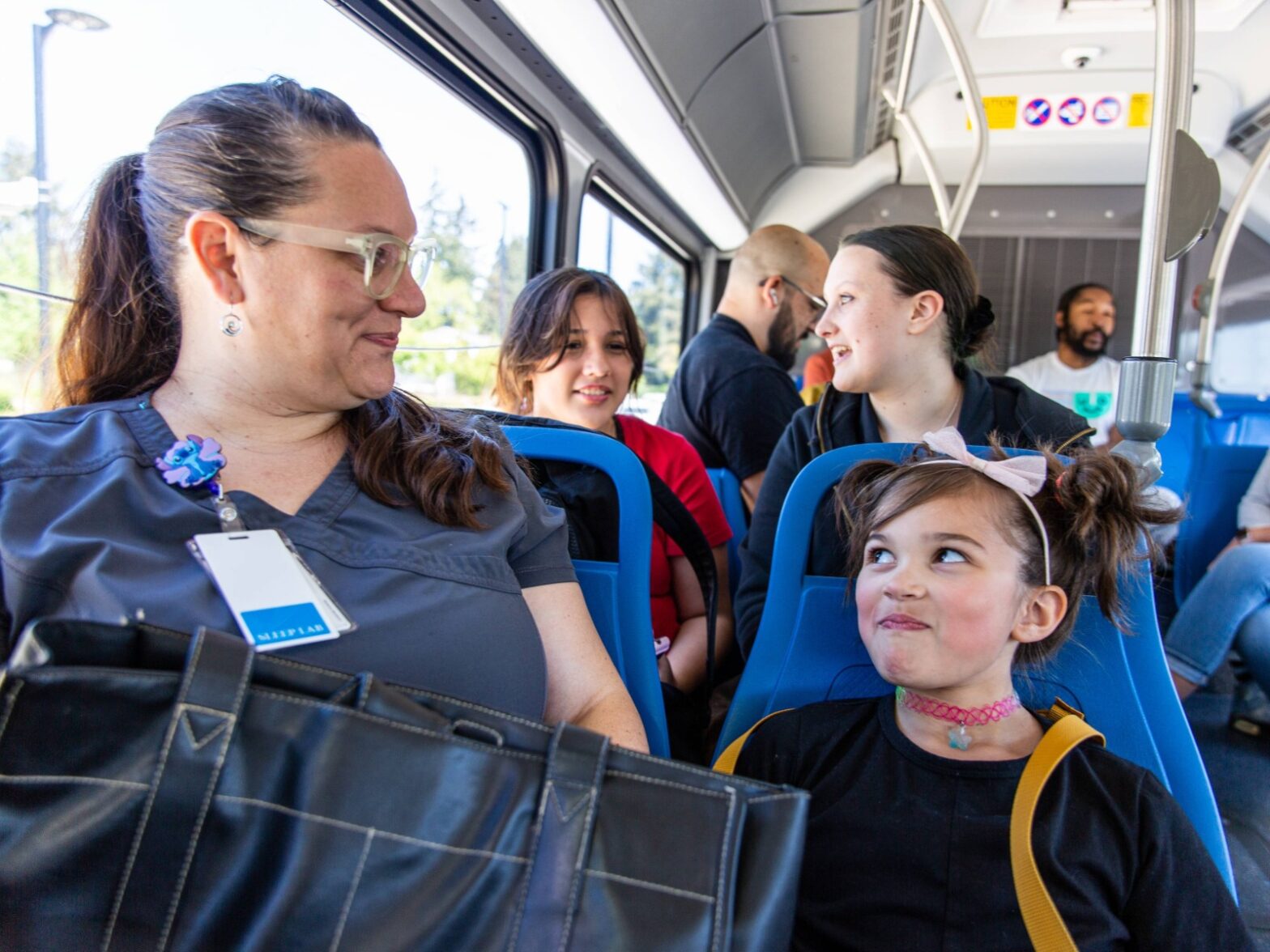 A woman in scrubs sits on a bus next to a young girl with a bow headband. Other passengers are seated behind them, engaged in conversation.