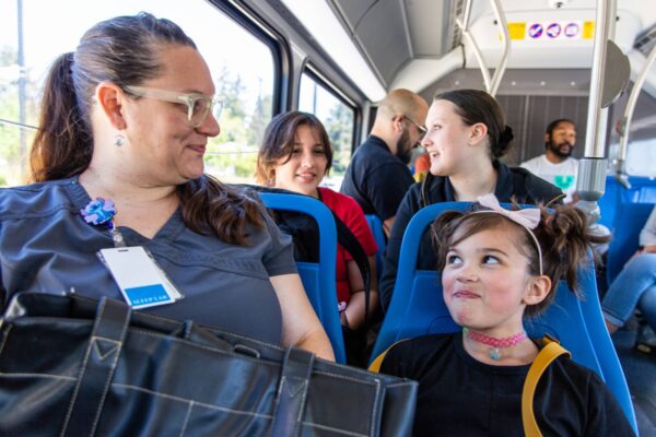 A woman in scrubs sits on a bus next to a young girl with a bow headband. Other passengers are seated behind them, engaged in conversation.
