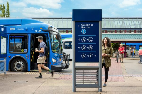 Two people are near a public transit sign and blue bus at a bus terminal on a clear day; one person is walking, another is standing and smiling.