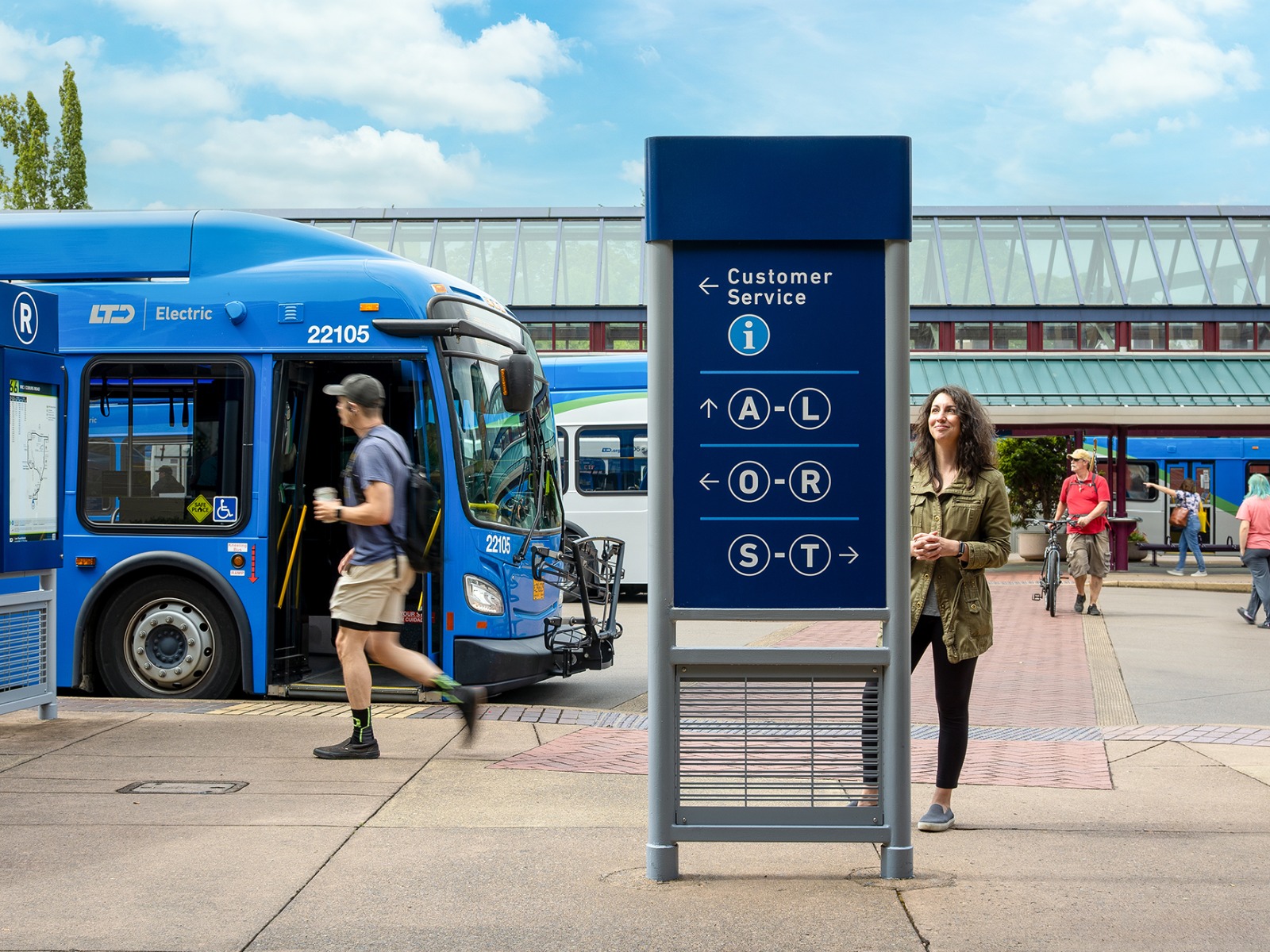 Two people are near a public transit sign and blue bus at a bus terminal on a clear day; one person is walking, another is standing and smiling.