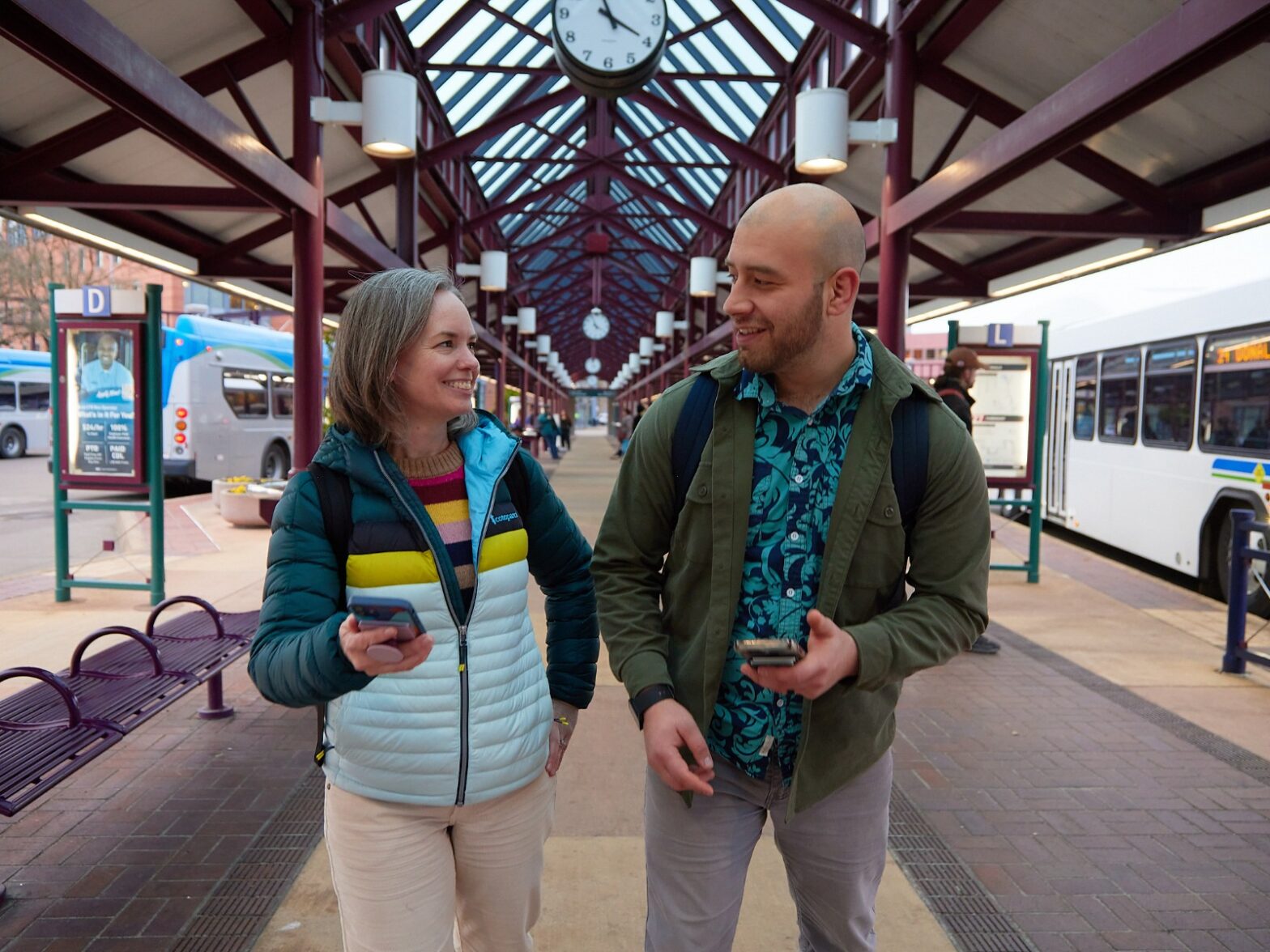 Two people walk and chat at a bus station, each holding a phone. Buses are parked nearby and a large clock hangs overhead.