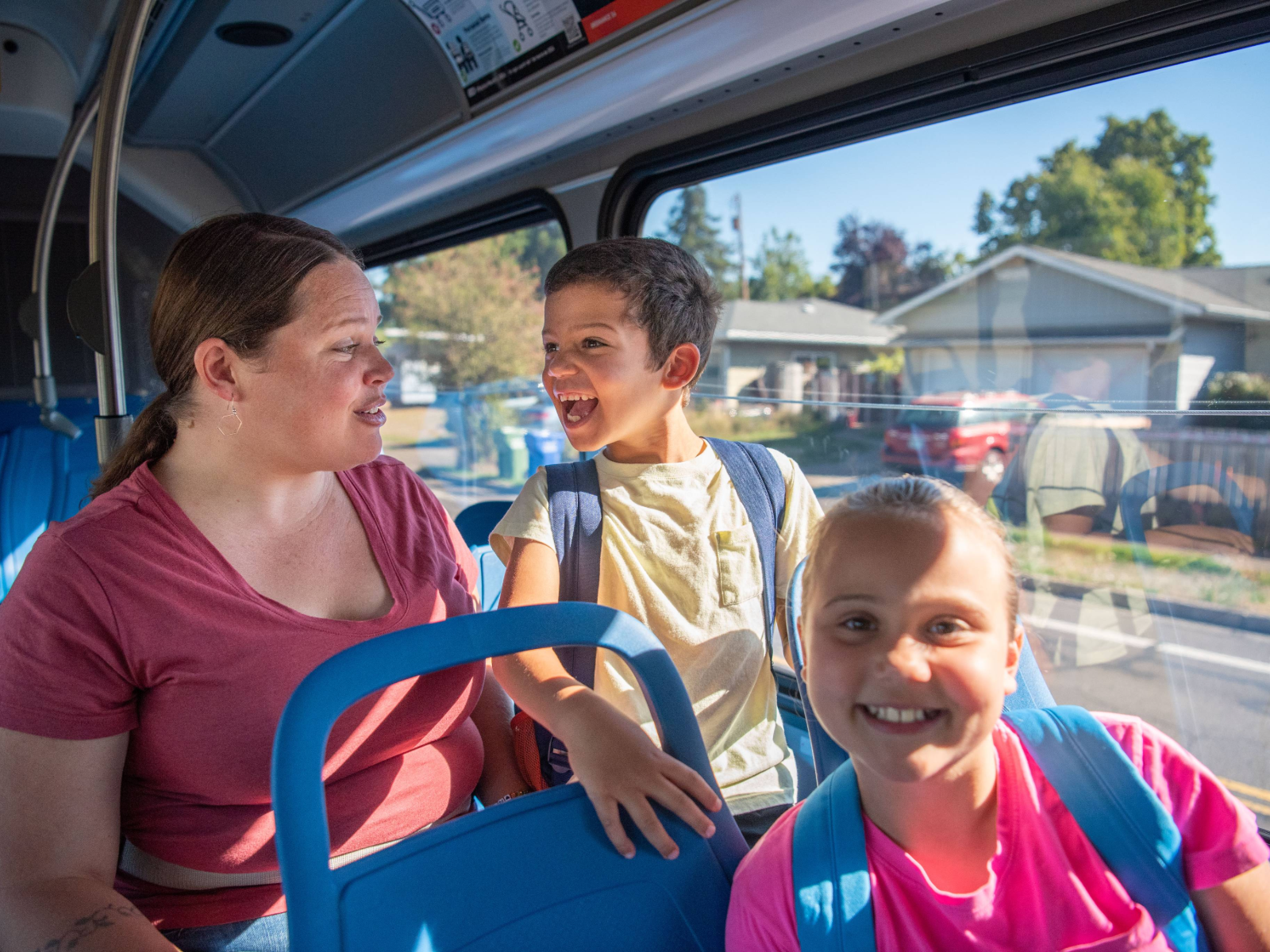 An adult and two children with backpacks are sitting together on a bus during the daytime; the children are smiling and looking cheerful.