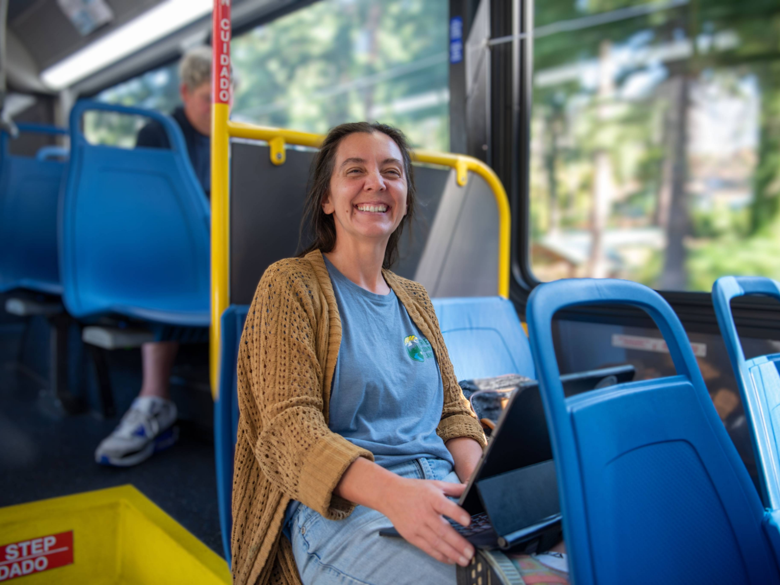 A woman wearing a blue shirt and brown cardigan sits smiling on a city bus, holding a tablet on her lap. Blue seats and a yellow safety pole are visible around her.