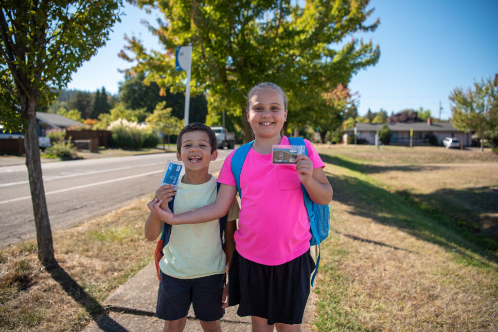 Two children with backpacks stand on a sidewalk, smiling and holding up cards, with houses, trees, and a clear sky in the background.
